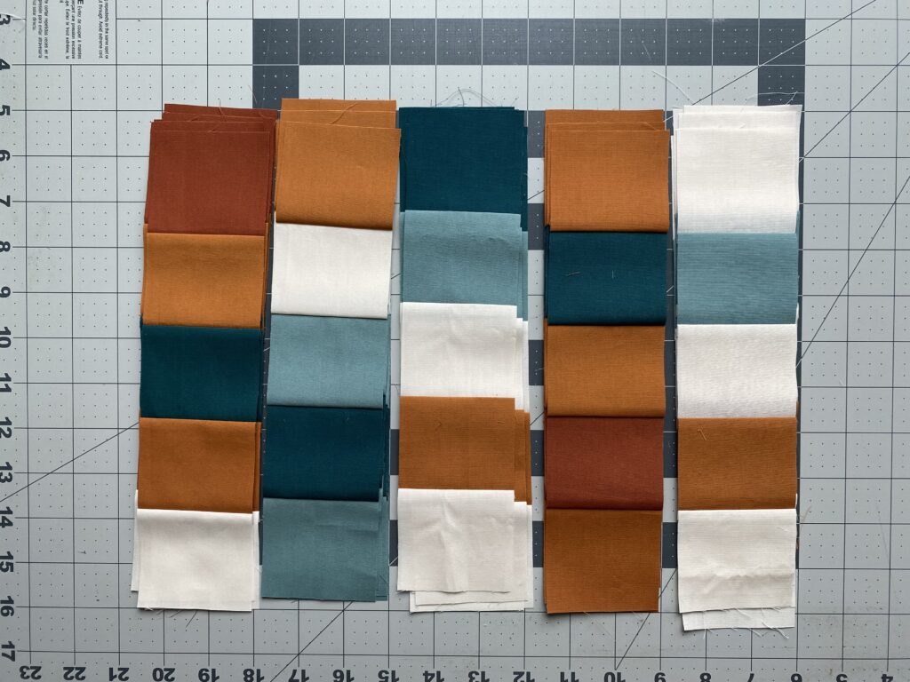 Rows of quilt blocks with seams pressed to the side in alternating directions, arranged by color on a cutting mat, prepared for sewing and nesting seams.
